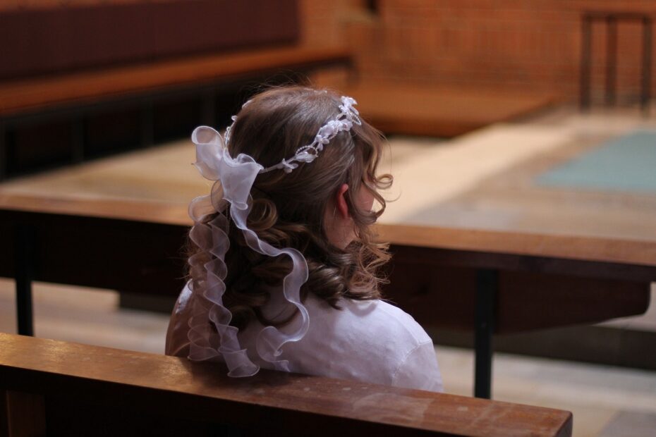 Das Bild zeigt ein Mädchen in einer weißen Festkleidung, das andächtig in einer Kirche sitzt. Ihr Haar ist kunstvoll gelockt und mit einem weißen Schleier sowie einer Blumenkrone geschmückt. Sie blickt nach vorne in Richtung Altar – ein Moment der Stille und Besinnung. Dieses Mädchen hat sich einen Spruch aus den 10 beliebtesten Konfirmationssprüchen ausgesucht. Ein Bibelvers, der sie auf ihrem Lebensweg begleiten wird, voller Hoffnung, Kraft und Glauben. Während sie auf den feierlichen Moment der Segnung wartet, denkt sie vielleicht über die Worte nach, die für sie nun eine besondere Bedeutung haben. Die warme Atmosphäre der Kirche, die Holzbänke und das gedämpfte Licht unterstreichen die Feierlichkeit dieses besonderen Tages – einem Meilenstein im Leben des Mädchens, das nun bewusst seinen Glauben bekennt.