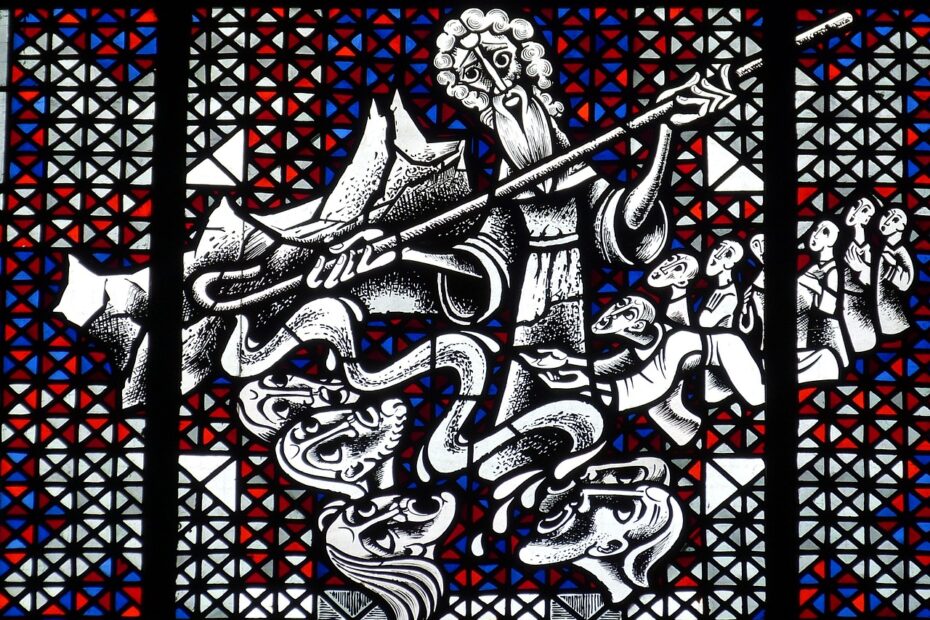 Das Bild zeigt ein kunstvolles Buntglasfenster mit einer biblischen Szene. Im Mittelpunkt steht eine zentrale Figur mit einem langen Bart, die einen Stab hält – dies ist der Stab von Mose. Die Szene scheint die Erzählung aus der Bibel darzustellen, in der Mose mit seinem Stab Wunder vollbringt. Im unteren Teil des Bildes sieht man schlangenähnliche Wesen und menschliche Köpfe, die möglicherweise den Moment symbolisieren, in dem Mose mit dem Stab von Gott gesandte Wunder vollbringt – zum Beispiel, als sein Stab sich in eine Schlange verwandelte oder als er das Rote Meer teilte. Im Hintergrund sind mehrere Menschen zu sehen, die offenbar ehrfürchtig oder erstaunt auf das Geschehen blicken. Das Farbenspiel des Buntglasfensters besteht aus geometrischen Mustern mit Rot-, Blau- und Weißtönen, was die Dramatik der Szene verstärkt.