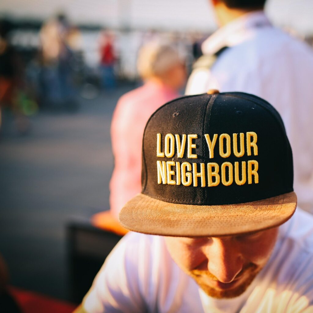 Das Bild zeigt einen Mann mit einer schwarzen Cap, auf der in großen, gelben Buchstaben der tiefgründige Spruch „LOVE YOUR NEIGHBOUR“ steht. Die Cap hat einen leicht abgenutzten Schirm, was ihr einen lässigen Look verleiht. Der Mann trägt ein weißes Shirt und blickt leicht nach unten, während der Hintergrund unscharf bleibt, mit Menschen in einer lebhaften Umgebung.

Dieser Moment erinnert daran, wie tiefgründige Sprüche kurz sein können und dennoch eine starke Botschaft vermitteln – in diesem Fall über Nächstenliebe und Zusammenhalt.