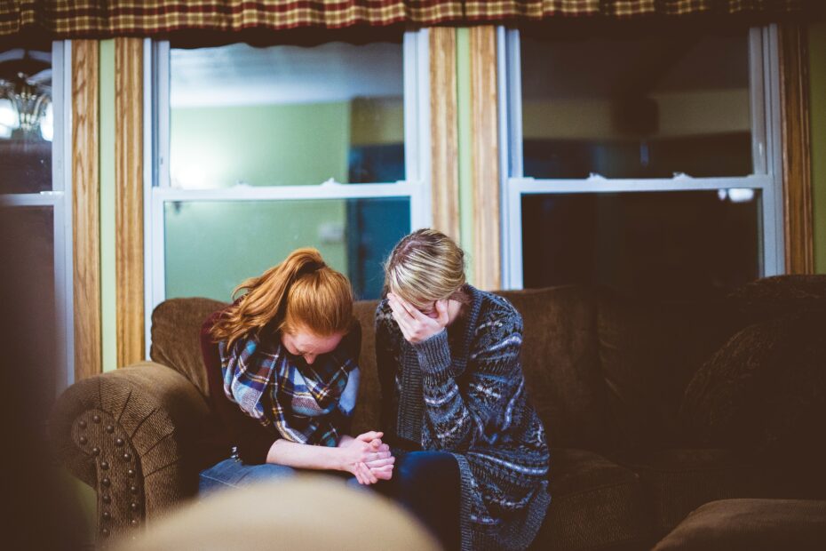 Das Bild zeigt zwei Frauen, die sich in einem Moment tiefer Trauer nahe sind und sich tröstende Worte zum Tod spenden. Während sie sich an den Händen halten, spiegelt ihre Körpersprache Mitgefühl, Schmerz und emotionale Unterstützung wider. Die Szene vermittelt eine Atmosphäre der Anteilnahme und zeigt, wie wichtig es ist, in schweren Zeiten füreinander da zu sein.